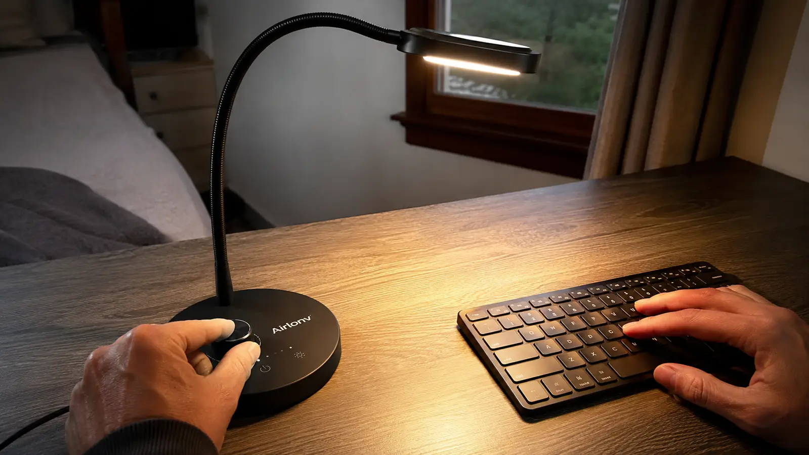 Hand adjusting the dimmer dial on the Airlonv LED desk lamp clamped to the edge of a home office desk in the evening