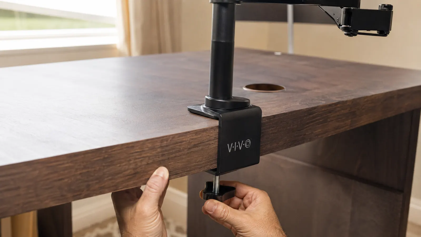 Close-up of hands clamping a monitor mount base to the back edge of a desk with a C-clamp