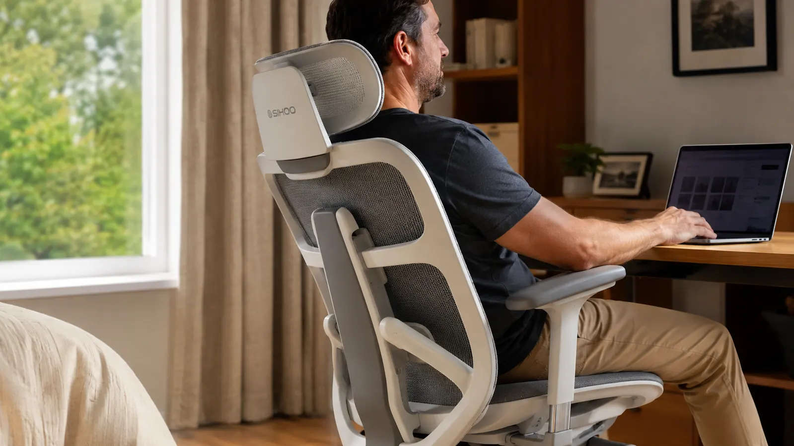 A man in a flannel shirt sitting at a home desk, leaning back into the mesh backrest of an office chair mid-workday